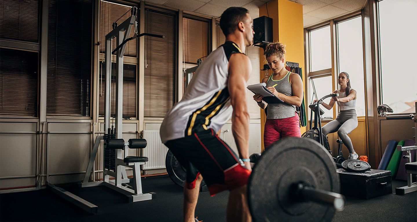 Personal trainer taking notes while a man performs a barbell deadlift in a gym